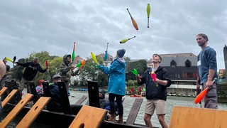 Ein Stocherkahn auf dem Neckar, der Himmel grau. Teilweise mit Kaputzen jonglieren junge Menschen dennoch mit bunten Kegeln. Zweiter von rechts: Weltmeister (2023) Moritz Rosner.