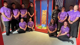 Mehrere Māori posieren um ein sogenanntes Pou im Museum der Universität Tübingen. Das Schnitzwerk ist kulturell und religiös sehr bedeutend.