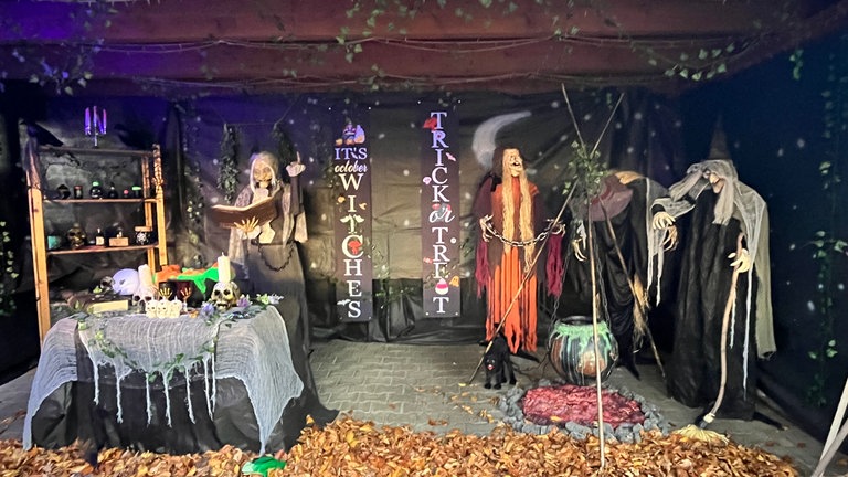 Vor einem Haus in Reutlingen-Degerschlacht ist ein Hexenhaus aufgebaut. Drei Hexen sind an einer Feuerstelle zu sehen, im Regal stehen Totenköpfe. 