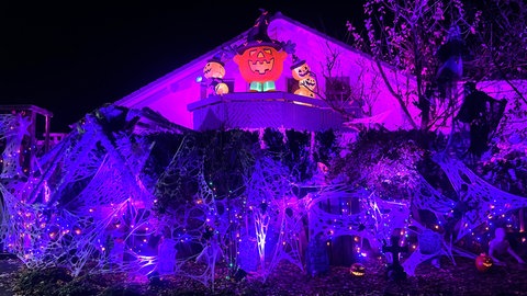 Ein Haus ist mit Halloween-Dekorationen geschmückt. Spinnweben, Grabsteine, Kürbisse und Lichteffekte sind zu sehen.