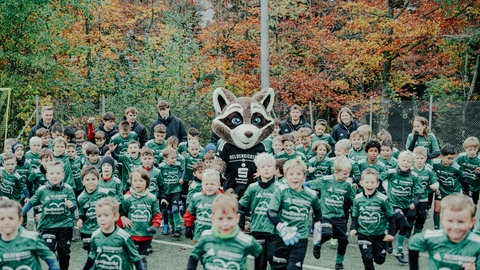 Kinder in grünen Trikots rechts und links von einem Waschbären-Maskottchen vor herbstlich gefärbten Bäumen. Zum Abschluss des Trainingstags beim Fußballcamp rennen die jungen "Helden" gemeinsam mit dem Heldenkicker-Maskottchen Raccoon über den Fußballplatz in Dietersweiler.