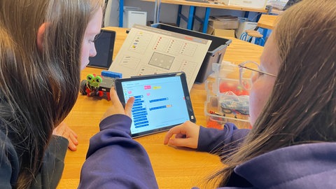 Zwei Schülerinnen tippen an der Realschule Horb auf dem Bildschirm eines Tablets. Blau hinterlegt sieht man eine lange Reihe von Befehlen, was der Roboter tun soll. Im Hintergrund der fertige Roboter von fischertechnik mit großen Augen.