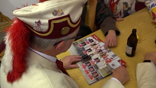 Der Zunftmeister von Rottenburg im Häs schaut ein Sticker-Album an. Die Zunft hat ein eigenes Sammelalbum mit allen Fasnetsfiguren. Seither ist Rottenburg im Sammelfieber.