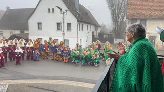 Die fast 91-jährige Lausbühlhexe Thea Breuling beobachter im Häs vom Balkon aus das Morgenwecken in Ergenzingen. Auf der Straße stehen einige Hexen, verkleidet mit Häs und Masken.