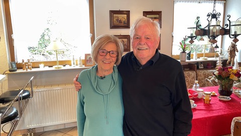 Auf dem Bild ist ein Älteres Ehepaar zu sehen. Die Frau (links) trägt einen türkisen Pullover, der Mann (links) trägt einen dunkelblauen Pullover. Beide lächeln.
