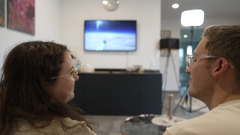 Ein Mann und eine Frau lächeln sich an. Im Hintergrund erkennt man einen Fernseher, auf dem der Film vom Verlobungsring in der Stratosphäre abgespielt wird.