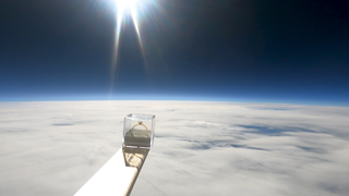 Eine durchsichtige Box mit einem goldenen Ring steht auf einer Holzlatte. Im Hintergrund erkennt man die Stratosphäre. Der Verlobungsring ist 30.000 Meter in die Höhe geflogen.