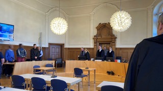 Im Gerichtssaal. Der Angeklagte in der Mitte hat sich erhoben. Links von ihm im blauen Pullover steht ein Dolmetscher, auf der anderen Seite der Verteidiger. Dem Angeklagten wird vorgeworfen, zwei Frauen mit einem Metallpoller in Horb am Neckar (Kreis Freudenstadt) angegriffen zu haben.  Dem Angeklagten wird vorgeworfen, zwei Frauen mit einem Metallpoller in Horb am Neckar (Kreis Freudenstadt) angegriffen zu haben. 