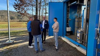 Unternehmer und Forschende unterhalten sich an der Forsthochschule Rottenburg vor dem blauen Container. In diesem befindet sich die Wasserstoffanlage.