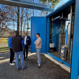 Unternehmer und Forschende unterhalten sich an der Forsthochschule Rottenburg vor dem blauen Container. In diesem befindet sich die Wasserstoffanlage.