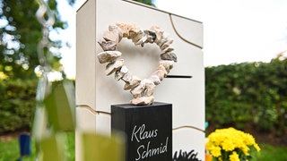 Ein Grabstein mit einem herzförmigen Kranz aus Steinen steht auf einem Friedhof. Oben hat er einen Briefschlitz. Innen ist er hohl. 