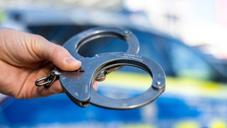 Eine Hand hält Handschellen vor einem Streifenwagen der Polizei. In Rottenburg wurde ein mutmaßlicher Schleuser festgenommen. (Symbolbild)