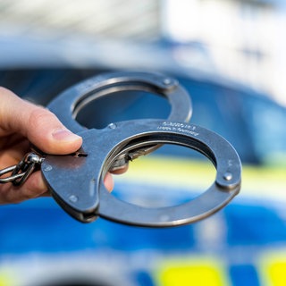 Eine Hand hält Handschellen vor einem Streifenwagen der Polizei. In Rottenburg wurde ein mutmaßlicher Schleuser festgenommen. (Symbolbild)