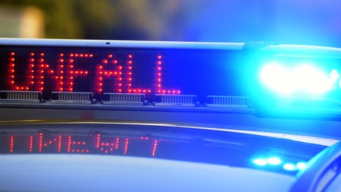 Symbolbild: Die blau leuchtenden Lichter auf einem Auto der Polizei und die digitale Aufschrift "UNFALL". Eine Frau wird eingeklemmt in ihre Autotür tot aufgefunden.