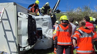 Lastwagen liegt kaputt auf einer Wiese neben einer Bundesstraße. Der Rettungsdienst steht um den LKW, der von der Straße abgekommen ist.