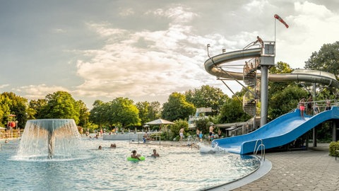 Ein blaues Becken im Schwimmbad mit einem Wasserpilz, einer blauen kleinen Rutsche und einer langen, kurvigen metallenen Rutsche, im Hintergrund Bäume und der Himmel ist bewölkt.