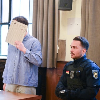 Der Angeklagte verdeckt sein Gesicht im Gerichtssaal. Er steht zwischen seinem Verteidiger und einem Justizvollzugsbeamten.
