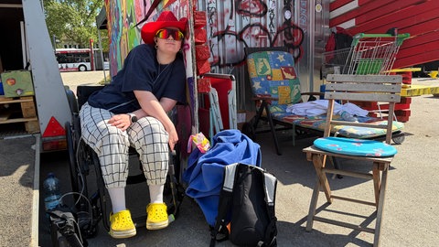 Anna-Lena Schmid sitzt am Rande der Liegenddemo in Tübingen. Seit zwei Jahren sitzt sie im Rollstuhl. Um sich vor äußeren Reizen zu schützen, trägt sie Sonnenbrille und Ohrenschutz. Wegen der Erschöpfung ist sie zeitweise auf Sauerstoff angewiesen.