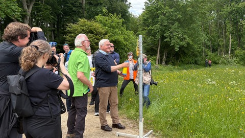 Steinmeier, in legerer Wanderkleidung, sprüht mit einer Sprühflasche auf einen Wegweiser. Viele Fotografen und Kameraleute haben ihre Objektive auf den Bundespräsidenten gerichtet.