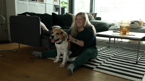 Simone Heusel sitzt in ihrem Wohnzimmer auf dem Boden und streichelt ihren Hund Emil, während er in ihrem Schoß sitzt. 