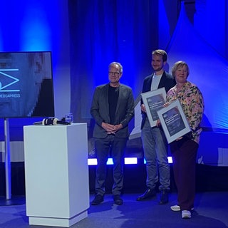 Die Beteiligten rund um ein Stehpult auf der Bühne, links die Moderatorin, rechts Intendant und Geehrte. Die Träger des Hans Bausch Mediapreises halten ihre Urkunden in die Kameras.