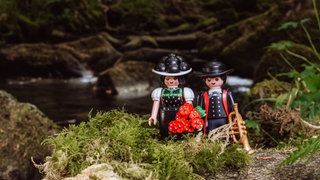 Die Playmobil-Figuren "Schwarzwald Marie" und Hannes in traditioneller Kleidung mit Bollenhut und Trompete in der Hand stehen im Wald.