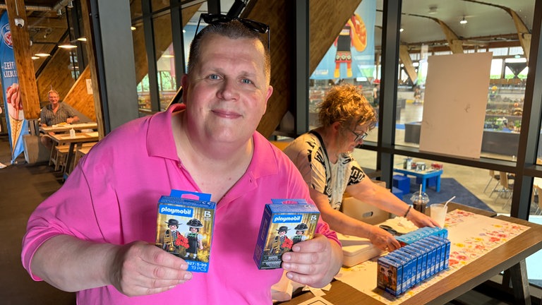 Ein Mann in pinkem Polohemd mit zwei Kartons mit den Spielzeugfiguren in den Händen.