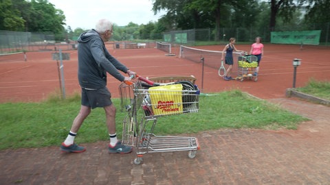 Tennistrainer Walter Betz schiebt sein Wägelchen mit Trainingsutensilien über den Platz.