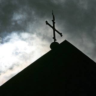 Im Gegenlicht und vor wolkenverhangenem Himmel ist eine Kirchturmspitze mit Kreuz zu sehen.