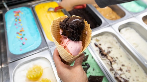 Buntes Eis in einer Auslage einer Eisdiele. Ein Verkäufer füllt gerade eine Waffel mit mehreren Kugeln. In einer Eisdiele in Ulm haben die Mitarbeiter an heißen Tagen viel zu tun. Nicht unbedingt ein Traumjob in der Hitze