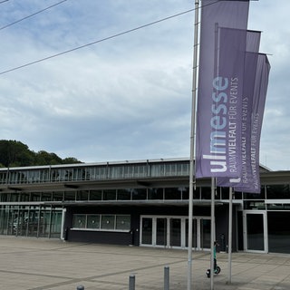 Der Eingang der Ulm Messe, ein flaches Gebäude mit mehreren Eingangstüren und Fahnen davor: In der Donauhalle der Ulm Messe will die AfD die Kandidatinnen und Kandidaten für die nächste Bundestagswahl bestimmen. Vor der Messe haben Organisationen wie auch Fraktionen des Gemeinderates in Ulm zu Gegendemos aufgerufen. Polizei und Stadt haben ein Sicherheitskonzept veröffentlicht.