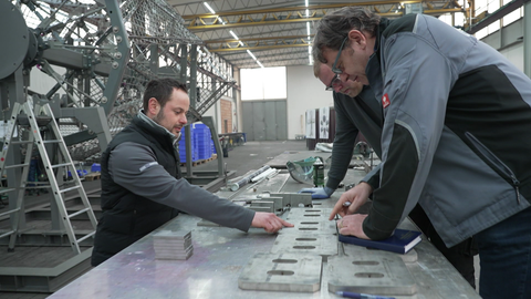 Drei Männer lehnen über einem Arbeitstisch in der Fabrikhalle des Gundelfinger Fassadenherstellers Josef Gartner GmbH. Sie arbeiten am Glaskreuz der Kathedrale Sagrada Família in Barcelona. Im Sommer 2026 soll das Bauwerk fertig sein.