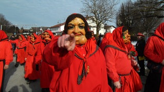 Eine in ein rotes Häs gehüllte Figur mit einer mit Kropf versehenen Frauenmaske aus Holz ist umringt zeigt mit dem Finger in die Kamera. Im Hintergrund sind mehrere Hästrägerinnen und -träger zu sehen.