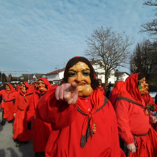 Eine in ein rotes Häs gehüllte Figur mit einer mit Kropf versehenen Frauenmaske aus Holz ist umringt zeigt mit dem Finger in die Kamera. Im Hintergrund sind mehrere Hästrägerinnen und -träger zu sehen.