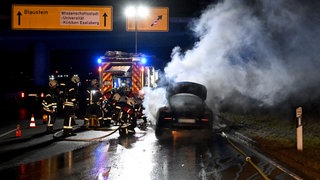 Ein qualmendes Auto auf der nächtlichen B10 bei Ulm. Mehrere Feuerwehrleute sind unter Flutlicht damit beschäftigt, das Auto zu löschen. Der Wagen hatte bei einer Probefahrt Feuer gefangen und brannte komplett aus.