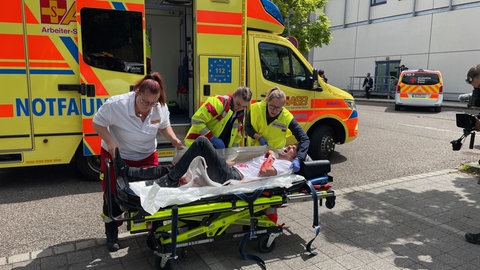 Ein Notfallwagen steht vor dem Blautalcenter in Ulm. Rettungskräfte kümmern sich um einen Mann auf einer Trage. Bei der Übung müssen sie unter anderem die laut Szenario schwerverletzten Menschen versorgen.