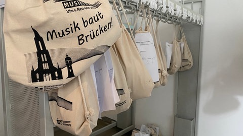 Mehrere Taschen, die für die Teilnehmenden des Deutschen Musikfestes in Ulm und Neu-Ulm bereit hängen. Auf den Taschen ist das Logo samt Motto "Musik baut Brücken" aufgedruckt. 