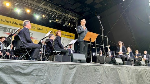 Ulms Oberbürgermeister Martin Ansbacher auf der Bühne auf dem Ulmer Münsterplatz. Er begrüßt die Besucherinnen und Besucher des Deutschen Musikfestes. Neben ihm die Musikerinnen und Musiker der beiden Stadtkapellen Ulm und Neu-Ulm.