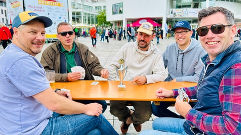 Eine Gruppe aus fünf Männern sitzen auf einer Bierbank auf dem Ulmer Münsterplatz und spielen während der Eröffnung des Deutschen Musikfestes das Karenspiel Binokel. Der Gewinner erhält einen Pokal.