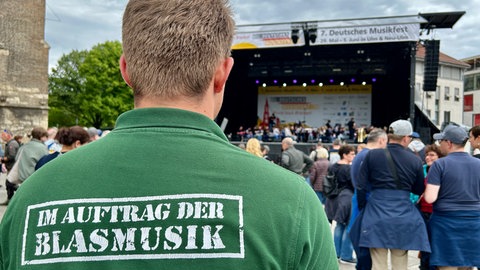 Ein junger Mann schaut auf die Bühne auf dem Ulmer Münsterplatz. Er trägt ein T-Shirt mit der Aufschrift "Im Auftrag der Blasmusik" auf dem Rücken.