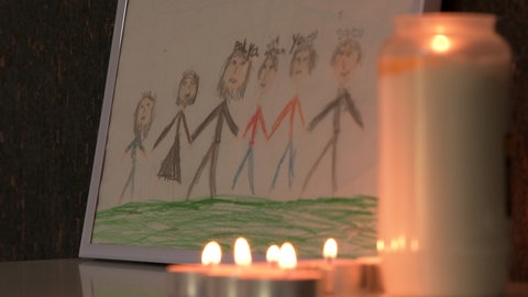 Teekerzen und eine Grabkerze brennen vor einem gemalten Bild mit Strichmännchen, die die Mitglieder der Familie zeigen. Gemalt hat das Bild Lana. Das Mädchen ist im August 2024 bei einem Unfall in Blaustein getötet worden. Ein 85-Jähriger hatte das Kind mit dem Auto erfasst. 
