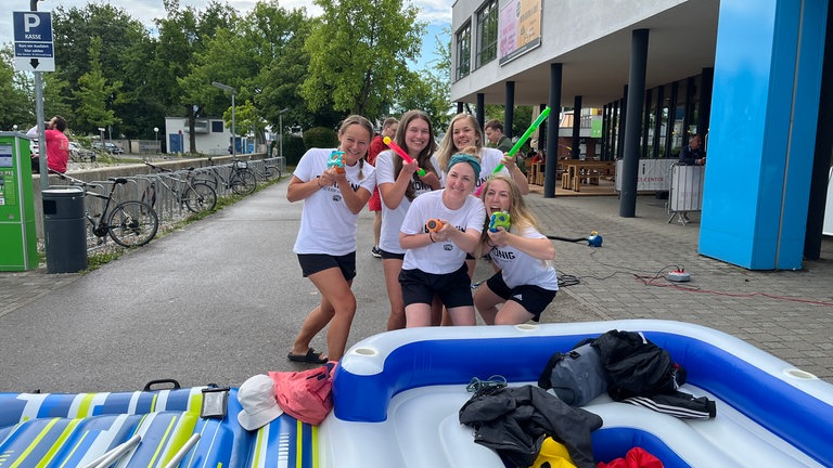 Fünf Frauen stehen hinter ihrem aufgeblasenen Gummiboot und zielen mit ihren Wasserspritzpistolen auf die Kamera.