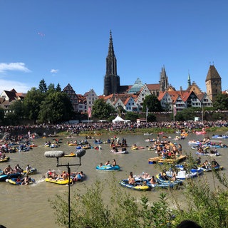 Viele Gummiboote und wilde Nabader auf der braunen Donau unter strahlendem Himmel.