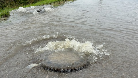 Aus Kanaldeckeln auf einer Straße sprudelt Wasser. Bei einem Gewitter mit Starkregen am Sonntagnachmittag ist die Kanalisation in Dornstadt (Alb-Donau-Kreis) überfordert. Die Wassermassen drücken Kanaldeckel hoch. Die Autobahn 8 ist kurzzeitig überflutet und muss für eine halbe Stunde gesperrt werden.