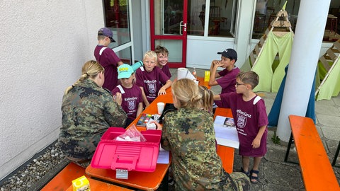 Soldatinnen sitzen in Uniform mit Kindern auf einer Bierbankgarnitur zusammen. Friedensaktivisten und -akivistinnen hatten in Kellmünz gegen ein Ferienlager der Bundeswehrn für Kinder protestiert.