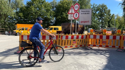 Ein Radfahrer steht vor einer Baustelle in Ulm mit einem großen Schild, auf dem "Donauradweg ab 01.08.2025 gesperrt" steht.