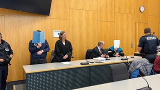 Angeklagte im Gericht verbergen ihre Gesichter mit Heftern: Das Landgericht Ulm hat im Prozess um Raubüberfälle auf ältere Frauen lange Haftstrafen gegen Vater und Sohn verhängt. 