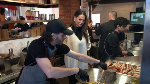 Mitten in der Küche der Currywurst-Bude. Die Geschäftsführerin und Mitarbeiter stehen auf dem engen Raum. Rechts werden Würste gebraten. Es ist viel los auf engem Raum.