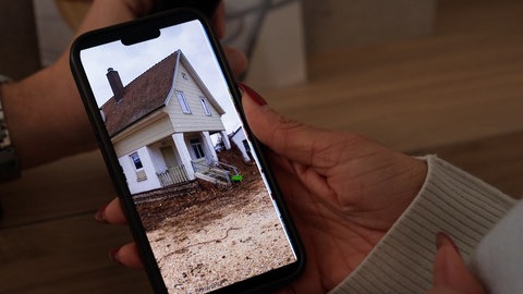 Ein Frau hat ein Handy in der Hand. Auf dem Handy ist ein Bild des alten Bahnwärterhäuschens. So hat es vor der Renovierung ausgesehen.