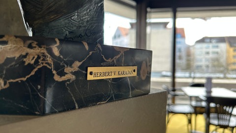 Nahaufnahme von dem unteren Teil der Büste, die Karajan darstellt mit einem Schriftzug. Die Büste steht im Theater Ulm auf einem Sockel, an dem ein Hinweisschild platziert werden könnte. 